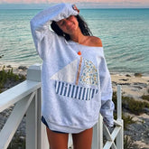 Woman wearing a light blue off-shoulder sweater with a sailboat design by the beach.